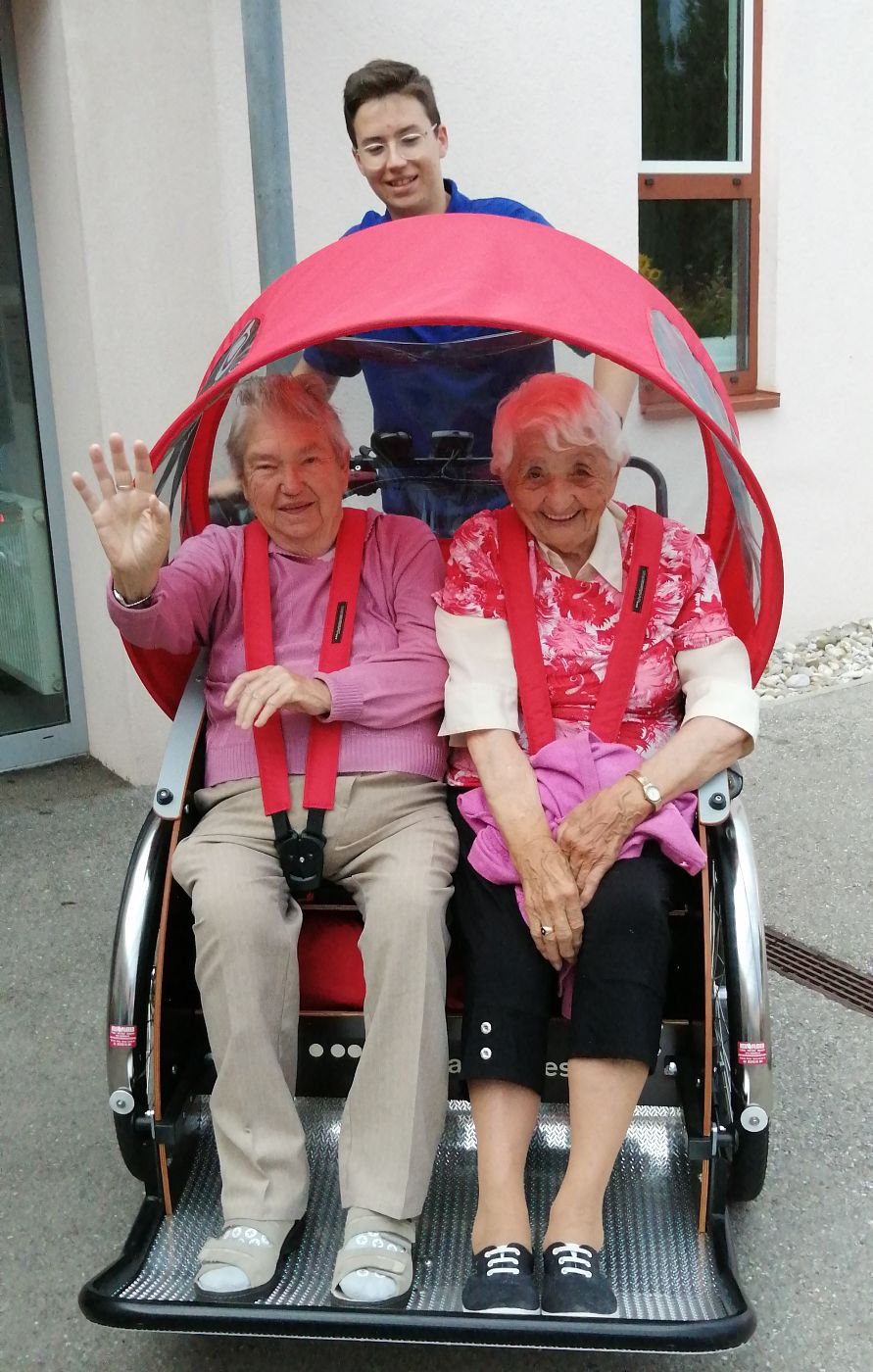 Ein älterer Herr und eine ältere Dame sitzen in einem Gefährt für 2 Personen, das von einem Zivildiener geschoben wird